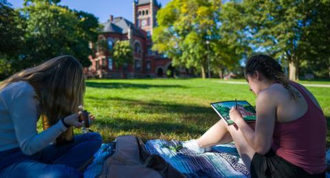 Two students sitting outside, one is drawing and the other is playing guitar